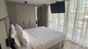 Ein modernes Hotelzimmer mit einem großen Bett, einem an der Wand befestigten Fernseher und Blick auf die Straße durch transparente Vorhänge.