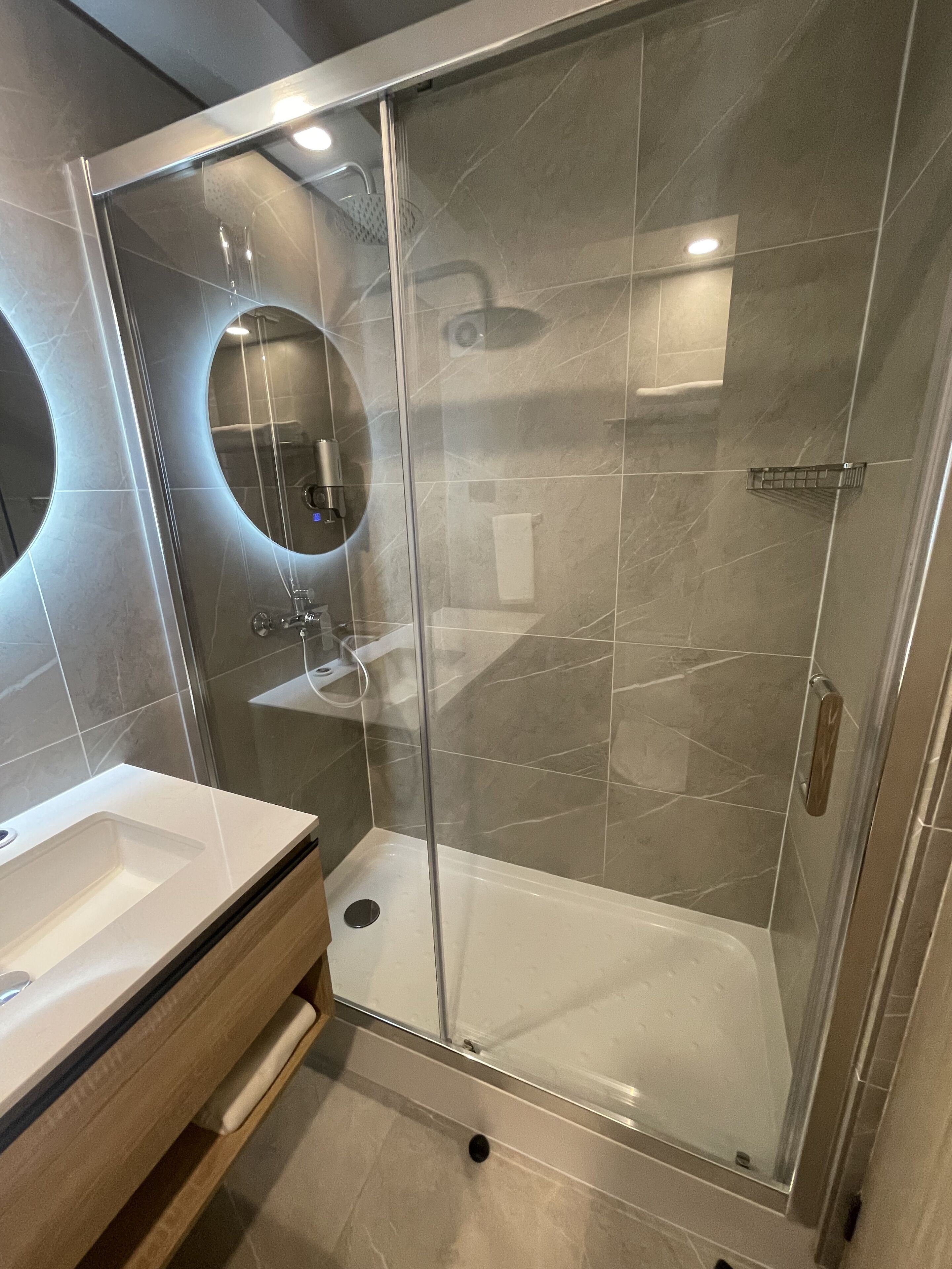 Een moderne badkamer met een douchecabine van glas, een ronde spiegel en een witte wastafel.
