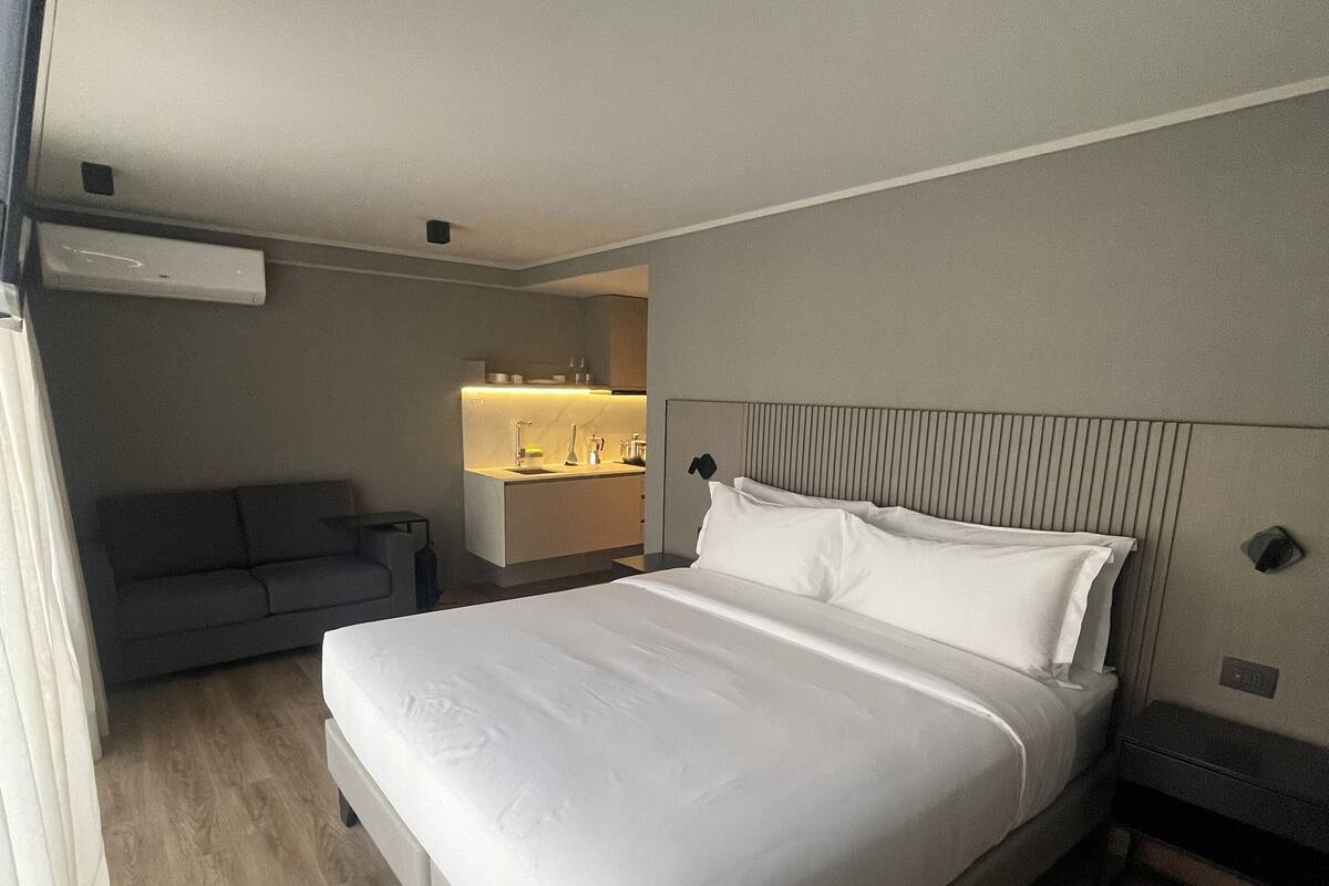 A modern hotel room with a large bed, a sofa, a small kitchenette, and a bathroom.