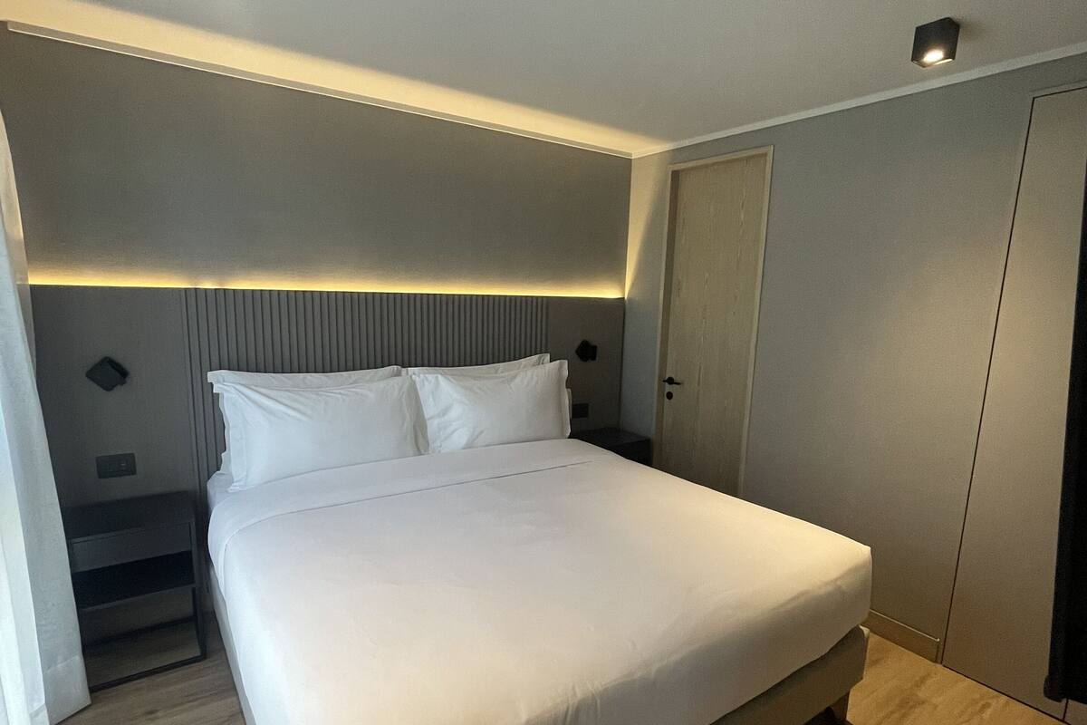 A modern hotel room with a large bed, a bedside table, a wardrobe, and a headboard with built-in lighting.