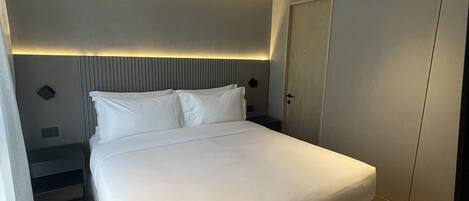 Una habitación de hotel moderna con una cama grande, una mesita de noche, un armario y un cabecero con iluminación integrada.