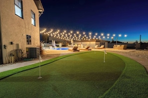 Un patio trasero bien iluminado con piscina, césped artificial y mobiliario de exterior.