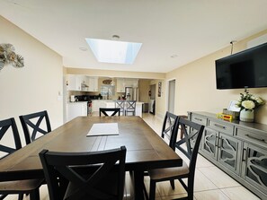 Un espace repas avec une longue table et des chaises, une cuisine à l’arrière-plan et un téléviseur à écran plat fixé au mur.