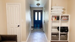 Un pasillo con una puerta a la izquierda, una escalera que sube a un cuarto oscuro y un área de almacenamiento con cestas y almohadones a la derecha.