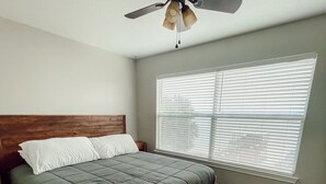 Un dormitorio con cabecera de madera, cama con colcha verde, ventilador de techo y ventana con persianas.