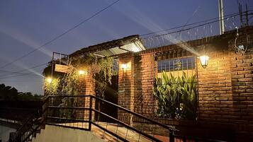 A brick building with a staircase leading to a covered area, surrounded by greenery and illuminated by wall-mounted lights.