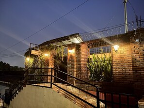 A brick building with a staircase leading to a covered area, surrounded by greenery and illuminated by wall-mounted lights. - Entre Tejas 1 (Concepción de Ataco)