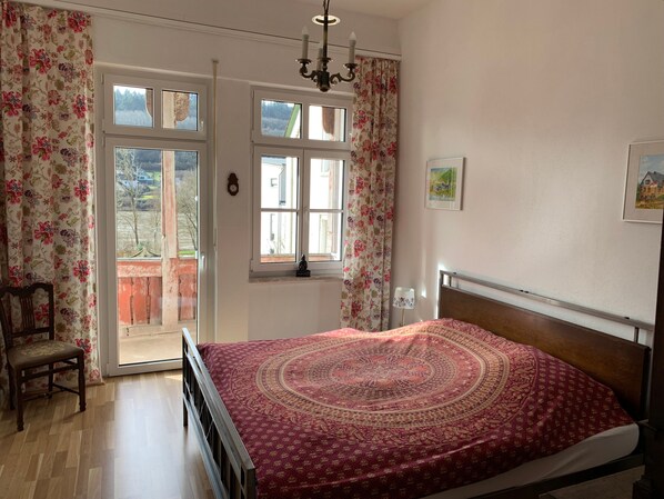 A bedroom with a bed, a chair, a chandelier, a window, and a door.