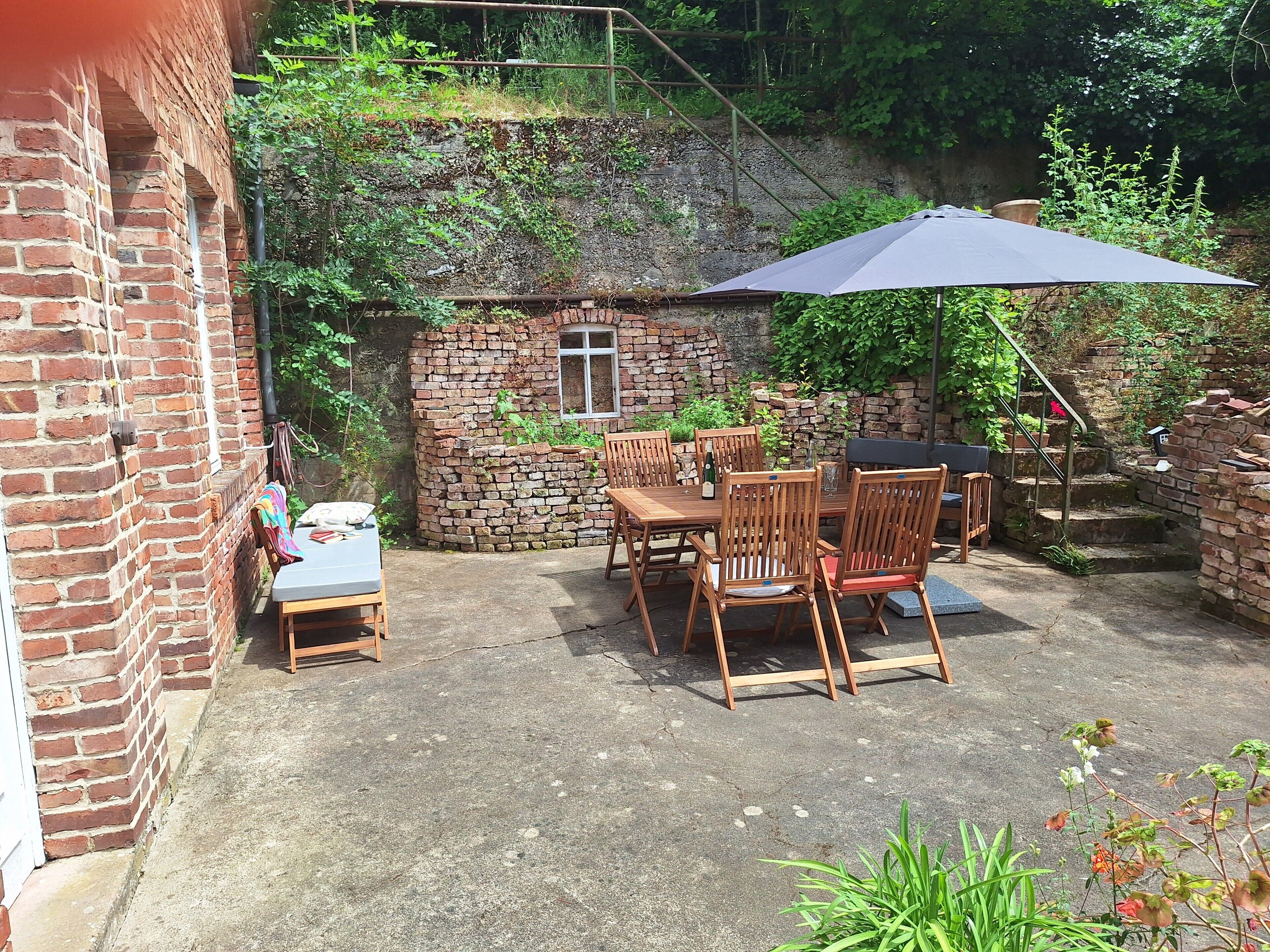 Eine Ziegelterrasse mit Tisch und Stühlen, umgeben von Grünpflanzen und einer Steinmauer.