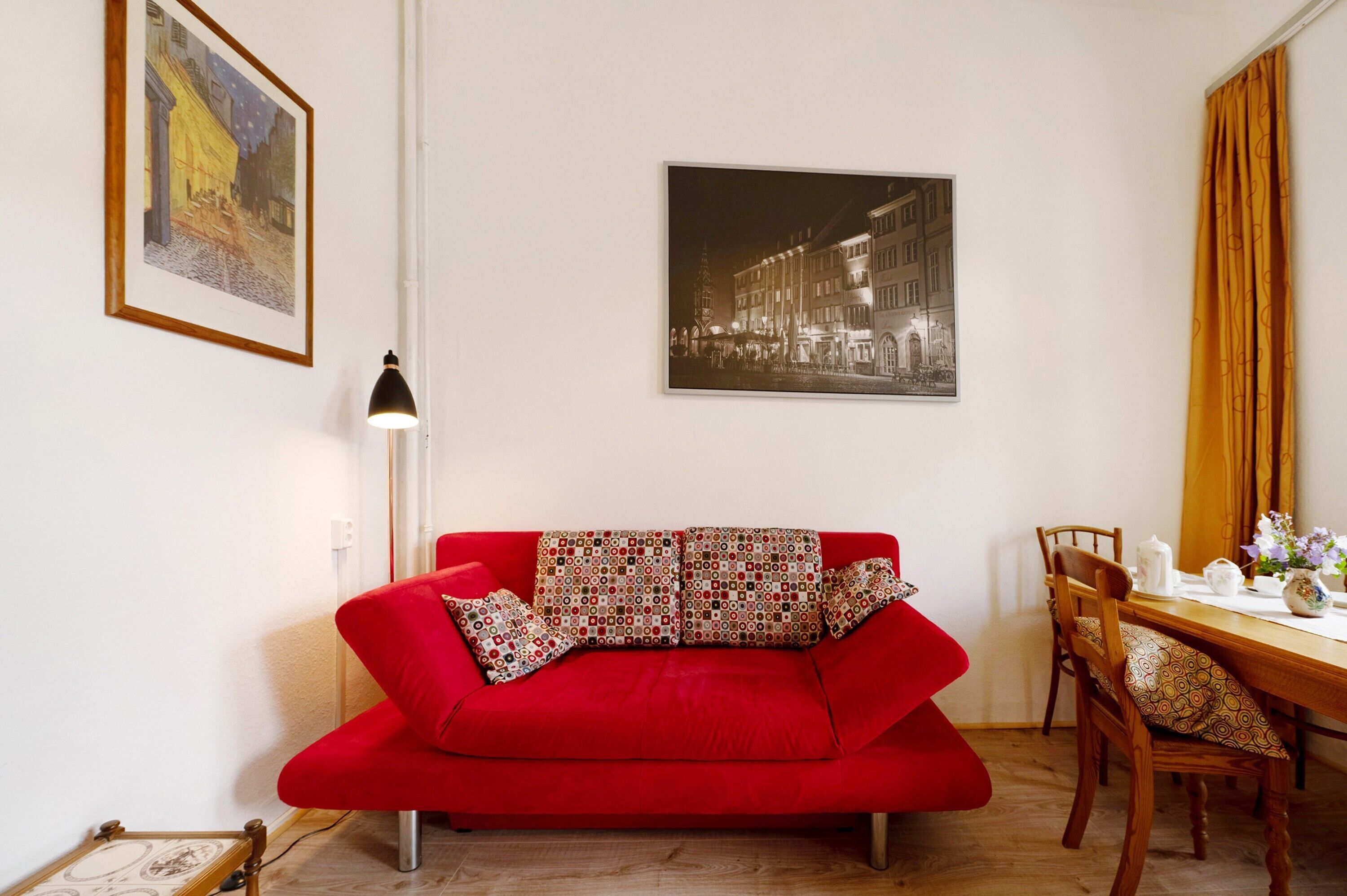 Ein rotes Sofa mit gemusterten Kissen, ein Esstisch mit Stühlen und zwei gerahmte Bilder an der Wand.