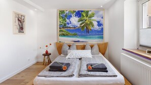 Ein Schlafzimmer mit Bett, Kissen, einem Bild mit Strandmotiv, einem kleinen Tisch mit Vase und einer Heizung.