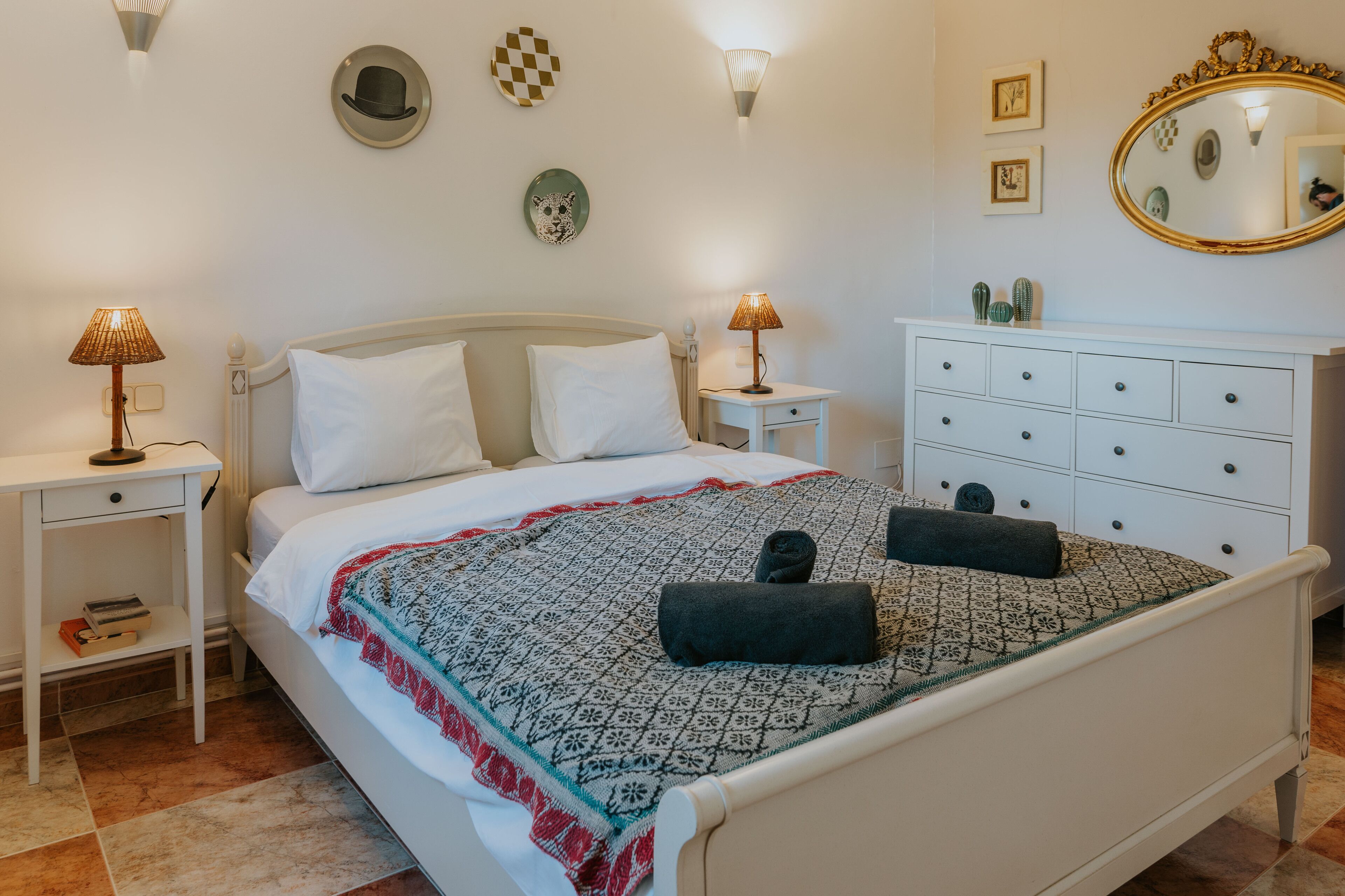A neatly made bed with a patterned blanket, two bedside lamps, a white dresser with drawers, and wall decor including a mirror and framed pictures.