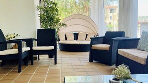 Ein sonnendurchflutetes Zimmer mit Korbmöbeln, einer großen Topfpflanze und Blick auf Gebäude durch große Fenster.