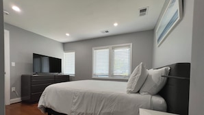 A bedroom with a bed, dresser, television, and windows with blinds.