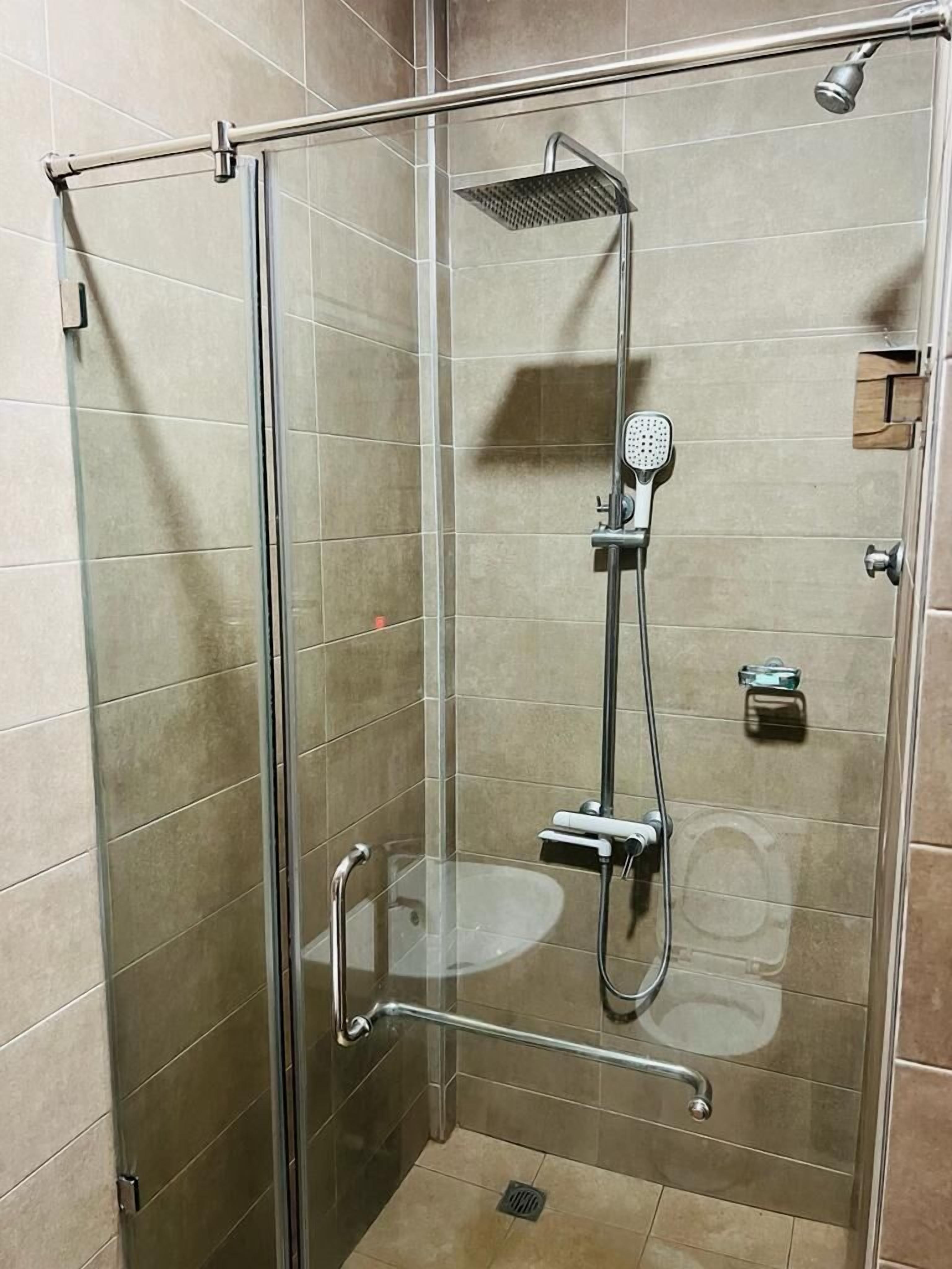A glass-enclosed shower stall with a handheld showerhead, a wall-mounted showerhead, and a glass door with a handle.