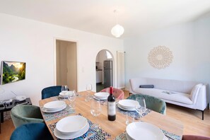 A dining area with a wooden table set for four, surrounded by chairs, a sofa, and a kitchen in the background.