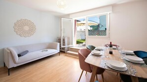 Una sala de estar moderna con un sofá, una mesa de comedor preparada para cuatro personas y un amplio ventanal con vistas a un jardín.