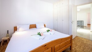 Una cama bien hecha con ropa blanca, un cabecero de madera y dos toallas con motivos de rosas. Hay un pequeño taburete con copas de vino y una botella. Se ve un armario blanco al fondo.