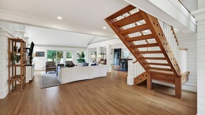 Un vaste espace de vie agrémenté d’un escalier en bois, d’un canapé blanc et d’une cuisine en arrière-plan.