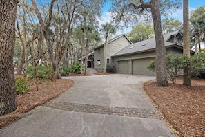 A residential driveway leading to a house with a garage, surrounded by trees and a landscaped area.