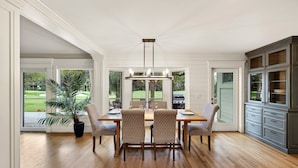 Un coin repas avec une table en bois, quatre chaises, un lustre et une vue sur un jardin grâce à de grandes fenêtres.