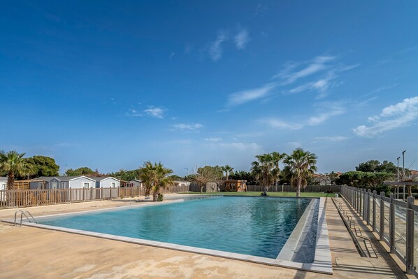 A rectangular swimming pool with a wooden deck and a fence, surrounded by palm trees and a clear blue sky.