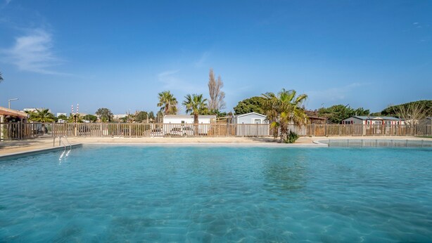 Una piscina cristallina blu con una recinzione in legno, palme e un edificio bianco sullo sfondo.