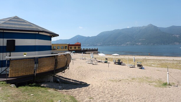 A beach with a building, beach chairs, umbrellas, and mountains in the background. - Mansarda Vialba Apartment in Holiday Home with Lake View and Balconies (Germignaga)