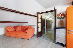 Une pièce avec un plafond orné de poutres en bois, un canapé orange, un meuble en bois et une porte donnant sur un couloir.