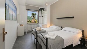 A hotel room with two beds, a small desk, a chair, a radiator, a window with a view of greenery, and a hanging light fixture.