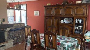 A dining area with a table set for four, surrounded by wooden chairs with cushioned seats. A wooden cabinet with glass doors is against the wall, displaying various dishes and kitchenware.