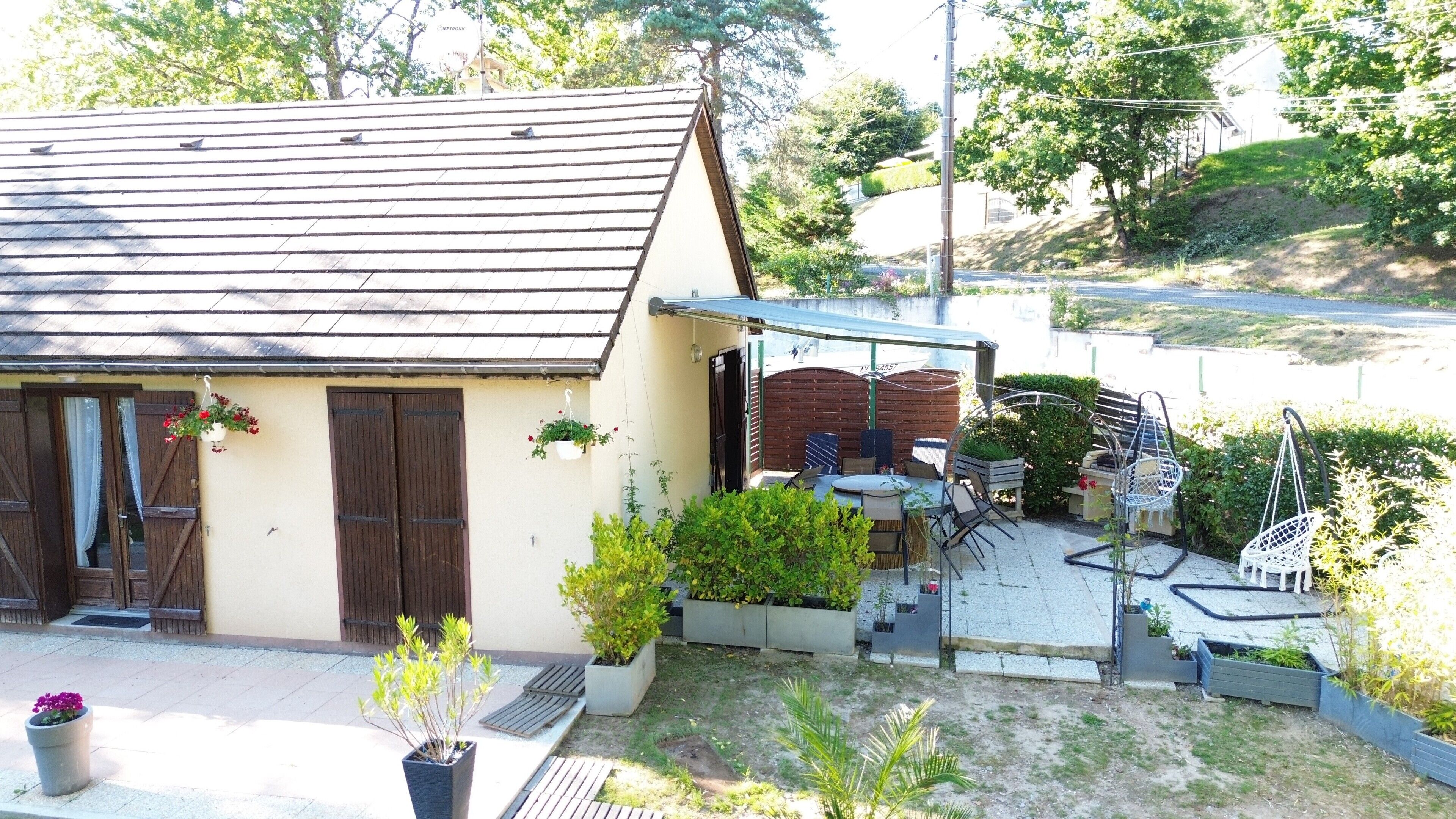 A house with a sloped roof, a patio with outdoor furniture, and a garden area with potted plants.
