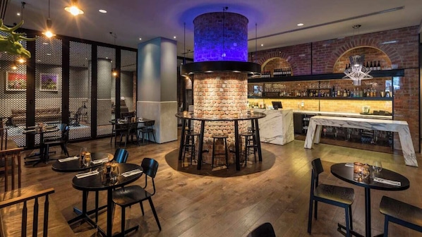 A restaurant with a central brick fireplace, wooden tables, and chairs. There is a bar area with shelves of bottles and a chandelier.