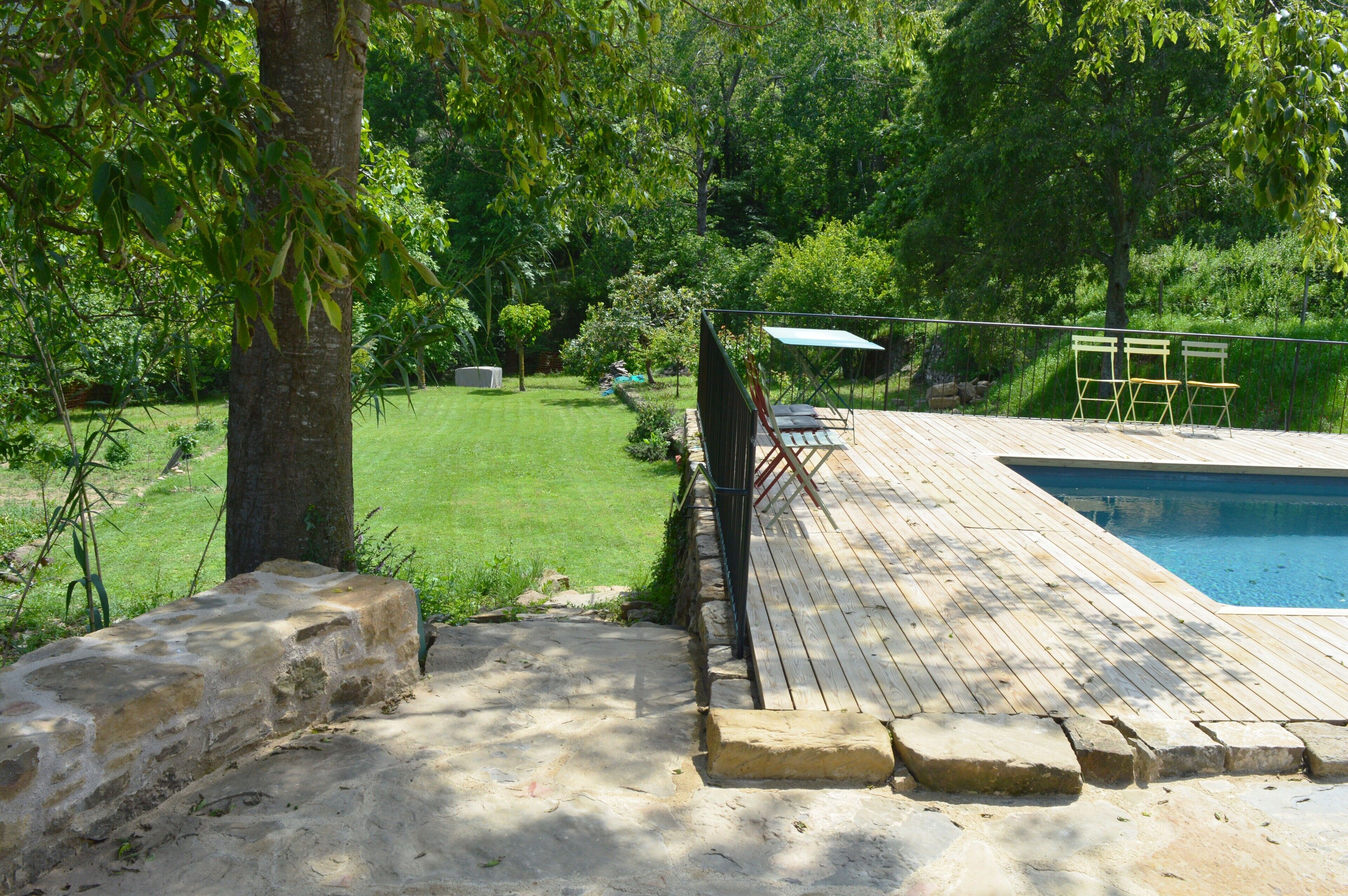 Un patio trasero con piscina, una terraza de madera y una zona ajardinada.
