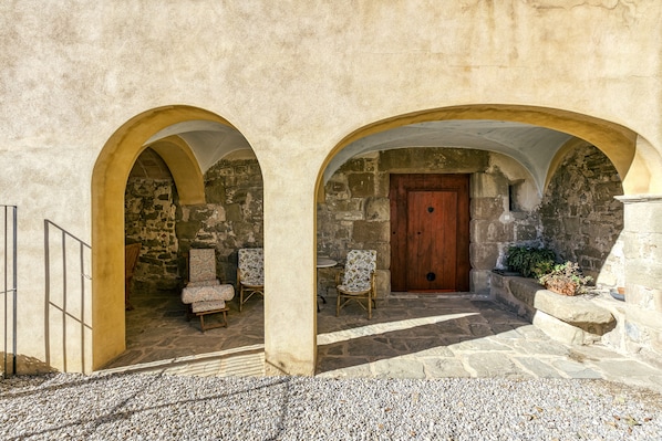 A stone building with two arched doorways, a wooden door, and two wicker chairs.