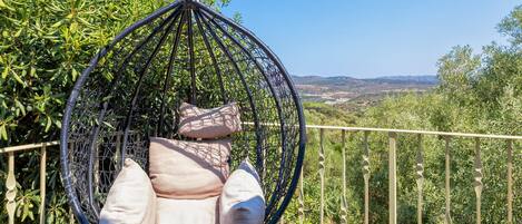 Una silla colgante con cojines, una valla de metal y vistas a zonas verdes y colinas.