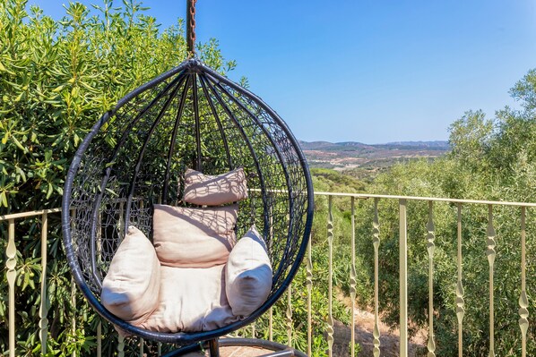 A hanging chair with cushions, a metal fence, and a view of greenery and hills.