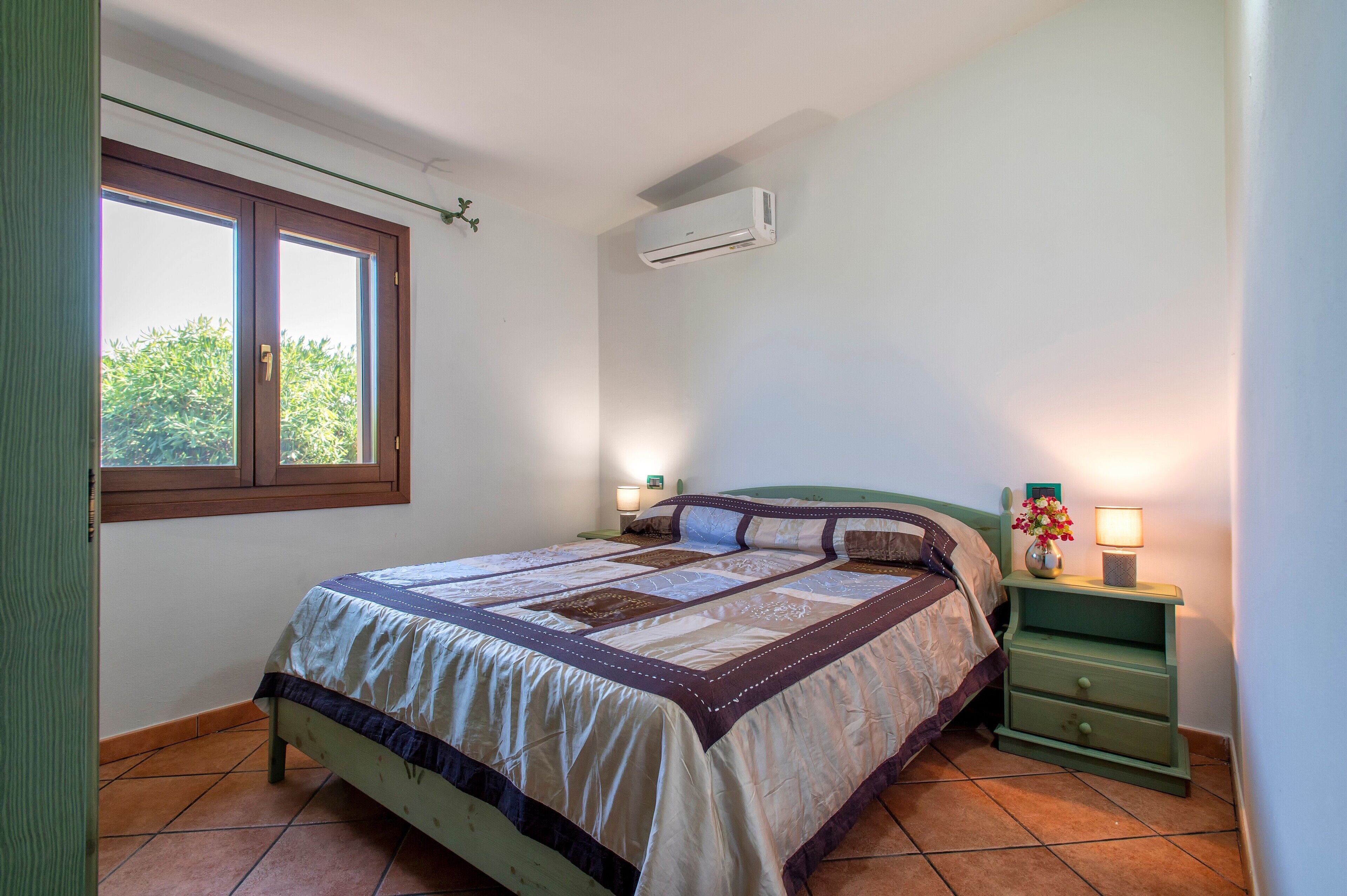 Ein Schlafzimmer mit Bett, Nachttisch, Wand-Klimaanlage, Fenster und Blick auf Grünfläche.