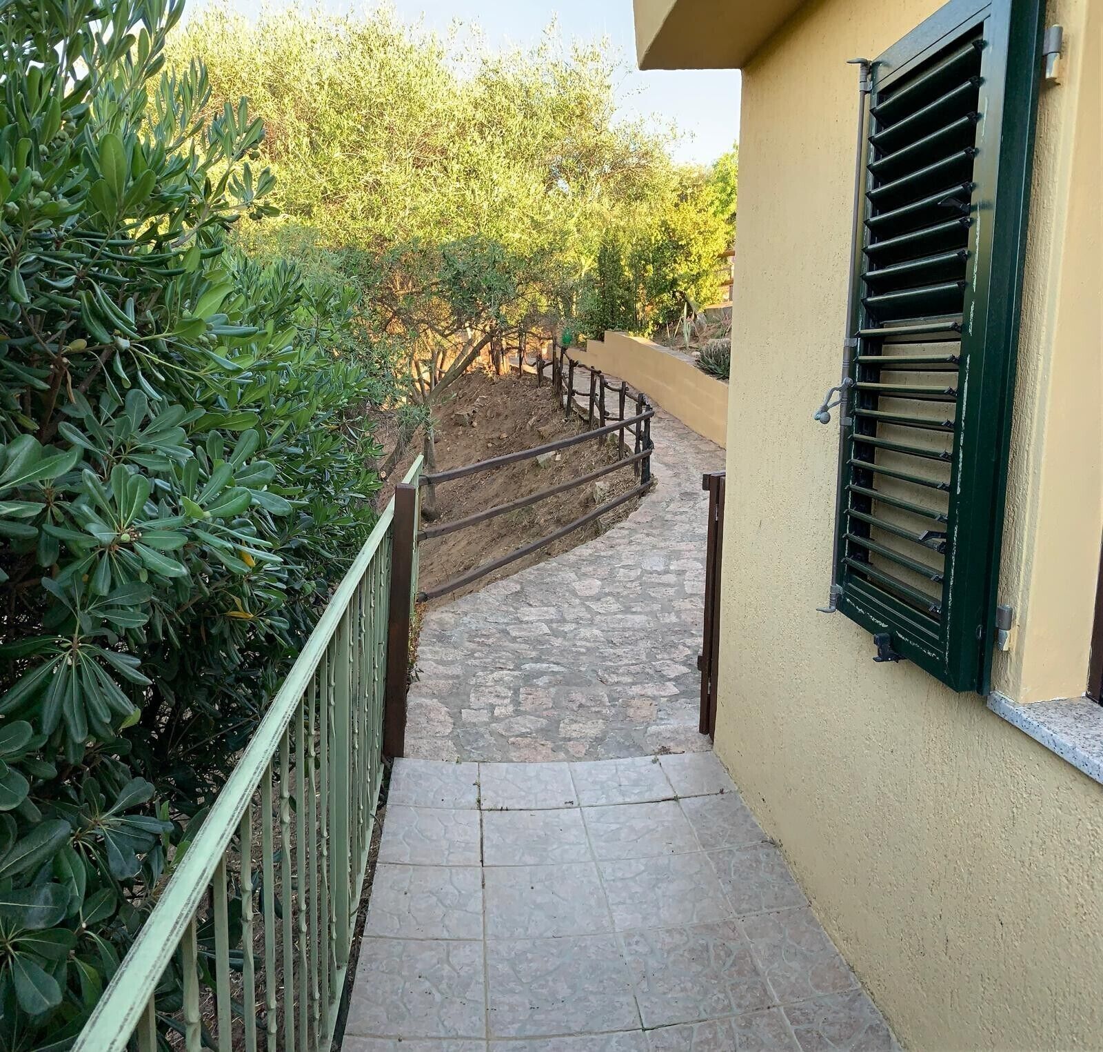 Un estrecho sendero de piedra que conduce a un edificio con barandilla verde y una ventana con persianas.