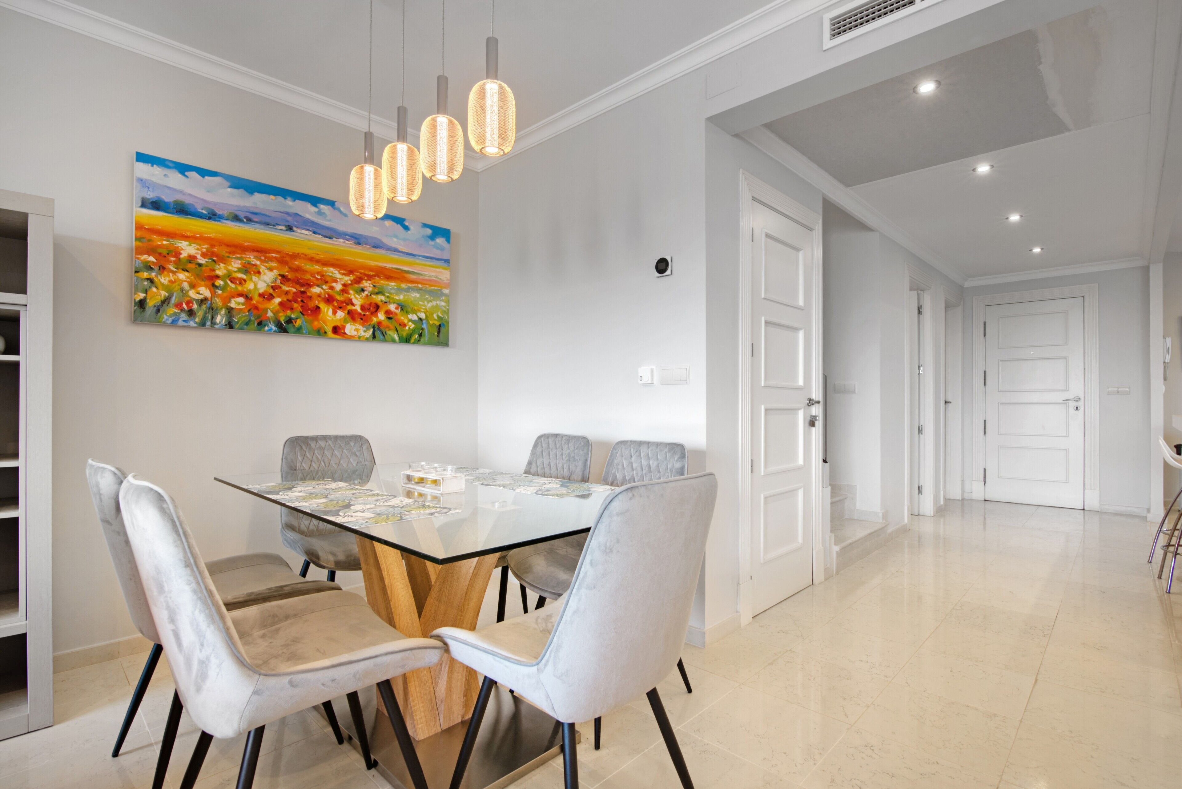 Un comedor con mesa de vidrio, sillas tapizadas de gris, iluminación colgante y un cuadro de paisaje grande en la pared.