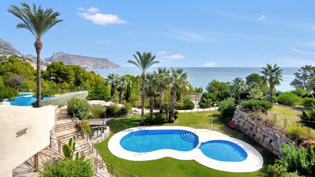 Una piscina dalla forma sinuosa, circondata da un muro in pietra e palme, con vista sul mare e sulle montagne sullo sfondo.