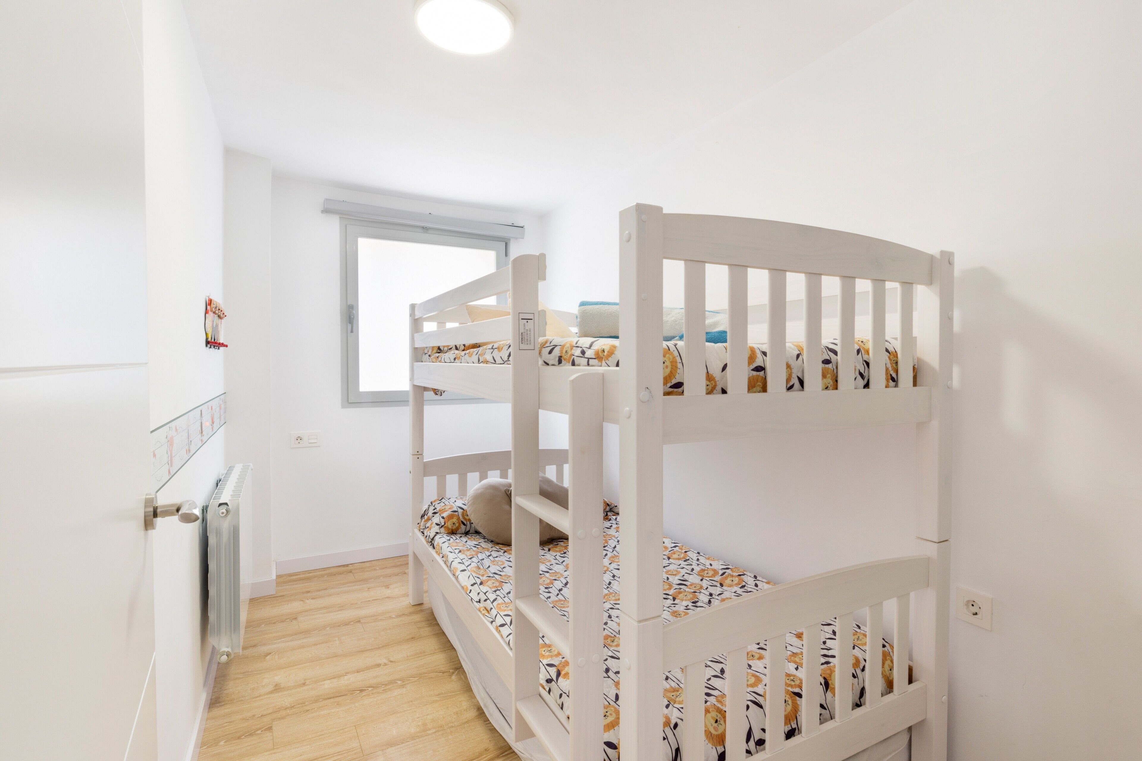 Een kinderkamer met stapelbedden, een radiator, een raam en een witte muur.