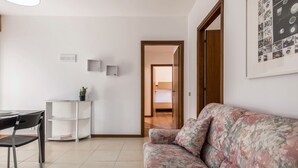 Quarto com sofá, escrivaninha, cadeira, uma planta pequena e porta que dá para outro cômodo.