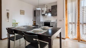 Uma sala de jantar com uma mesa posta para quatro, cadeiras, um lustre e uma cozinha ao fundo.