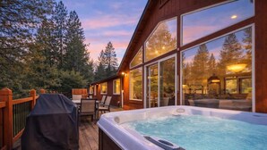 Un jacuzzi en una terraza de madera con vistas a un bosque y un edificio con ventanas grandes.