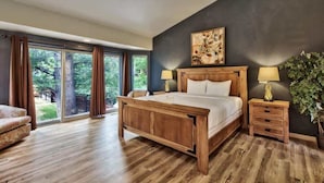 Un dormitorio con una cama grande, mesitas de noche de madera, un cuadro y vistas a través de una puerta corrediza de vidrio.