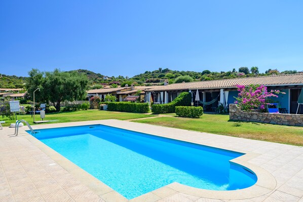 A swimming pool with a tiled deck, surrounded by a well-maintained lawn and a row of buildings with terracotta roofs.