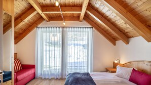 A room with a sloped wooden ceiling, a bed with white linens, a red sofa, and a window with sheer curtains.