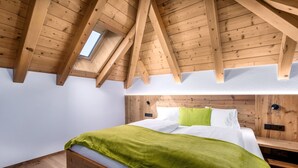 Un dormitorio con techo de madera inclinado, tragaluces, cama con ropa blanca y un plaid verde, cabecera y mesitas de noche de madera.
