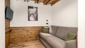 A corner room with a wooden bench, a grey sofa, and a wall-mounted TV. - Apartment "Perfila Dolomitenblick" with Mountain View, Shared Garden & Wi-Fi (Valdaora)